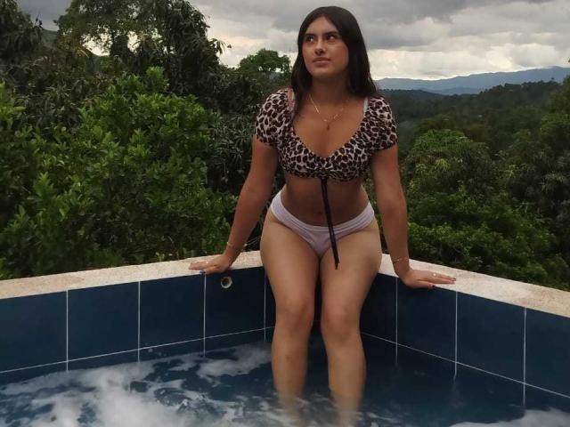 AnnieHotStar with brown hair, wearing a leopard print top and white panties, sits in a blue-tiled hot tub, water up to her thighs, looking off to the side.