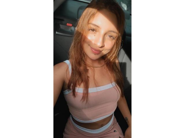 SaarahJones with dirty blonde hair, light pink ribbed crop top and matching pants, sitting in car, smiling, sunbeam on face, showing small tits and slim ass.