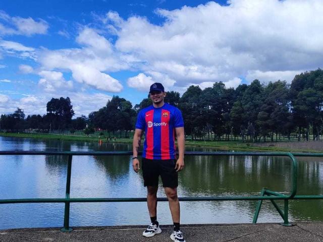 ErickStonee stands in front of a pond, wearing a blue and red striped sports shirt, black shorts, white sneakers, and a cap, with a tattoo on his right arm.