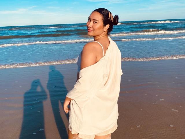 Ashlley69 è in piedi su una spiaggia, indossa una camicia bianca larga che lascia scoperta una spalla, i suoi capelli scuri sono raccolti in uno chignon, e guarda indietro, mentre le ombre di due persone si proiettano sulla sabbia..