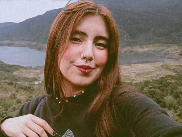 Diana Evans, avec des cheveux roux, un anneau de nez et des lèvres roses, souriante, portant une chemise noire, tenant un appareil photo, en extérieur, avec des montagnes et de l'eau en arrière-plan, légèrement flou..