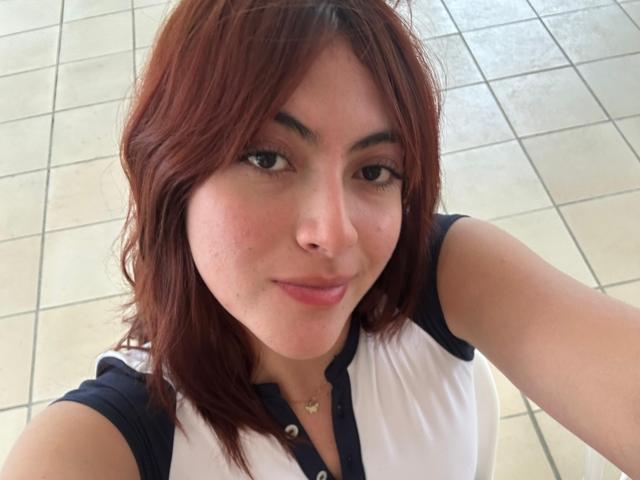 SpencerLe a la peau brun clair, des cheveux roux mi-longs, et elle sourit à l'appareil photo. Elle porte une chemise blanche et bleue, avec un petit collier en or..