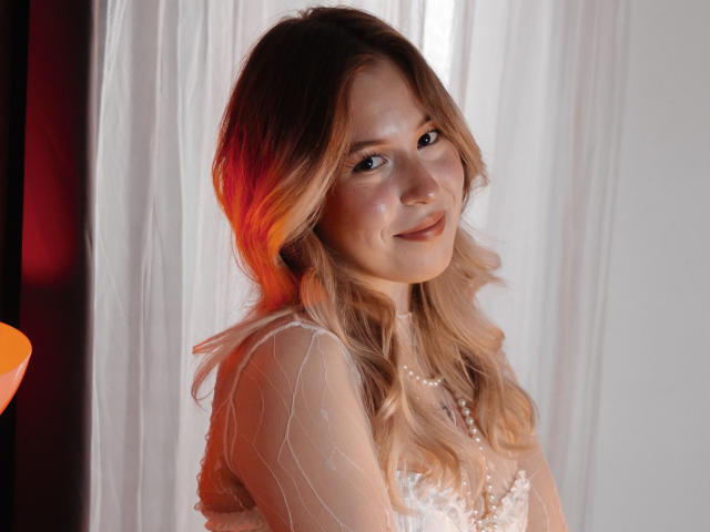 LanaRoselle, con i capelli biondi ondulati, indossa un abito bianco trasparente con motivi delicati, sorride dolcemente, con una leggera sfumatura rossa e arancione sul lato sinistro del viso..