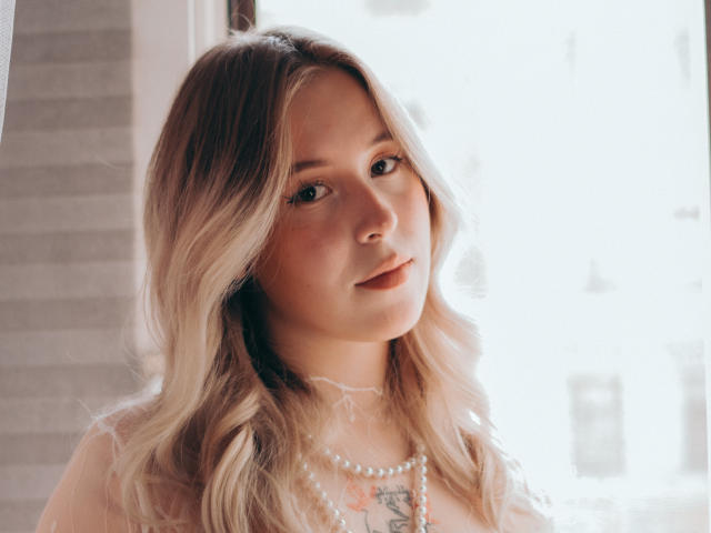 LanaRoselle a les cheveux blonds et ondulés, une peau claire et un tatouage discret sur la poitrine. Elle porte un collier de perles et regarde l'appareil photo avec un sourire doux..