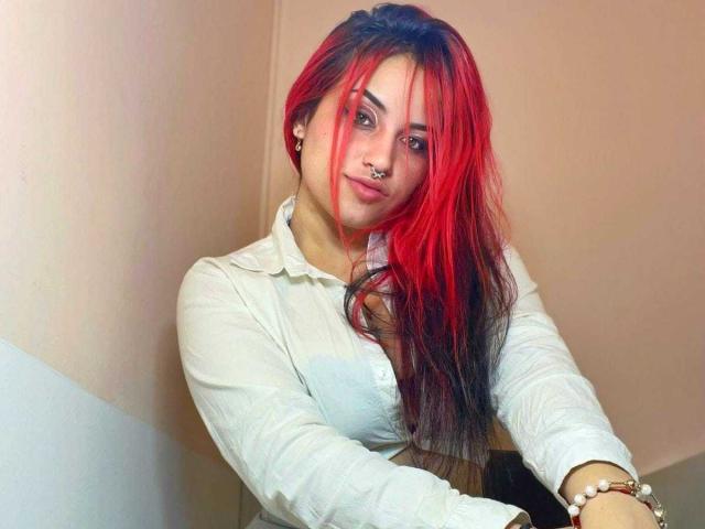 SophieRouge with red and black hair, nose ring, white shirt, and bracelet, smiling at camera, showing small tits, in a slightly dim room.