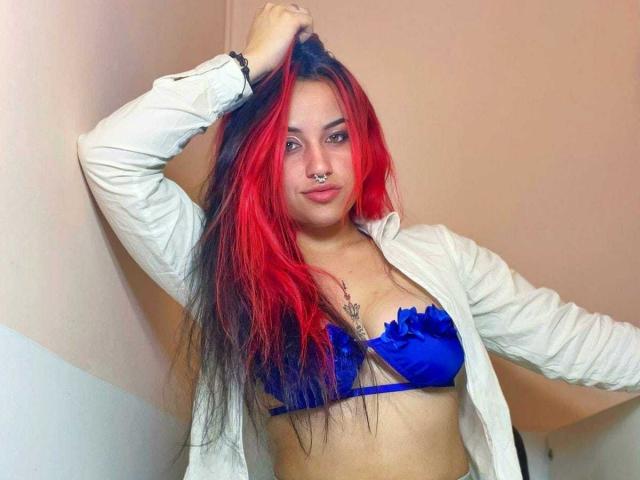 SophieRouge with red and black hair, nose ring, wearing a blue bra and white jacket, hand in hair, looking at camera, showing small tits and a slight smile.