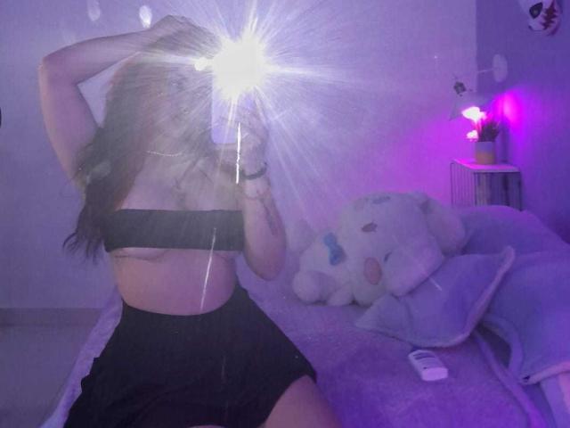 SophieRouge with long brown hair, black tube top exposing her tits, black shorts, taking a mirror selfie with a flash, purple bedroom, white stuffed animal on the bed.