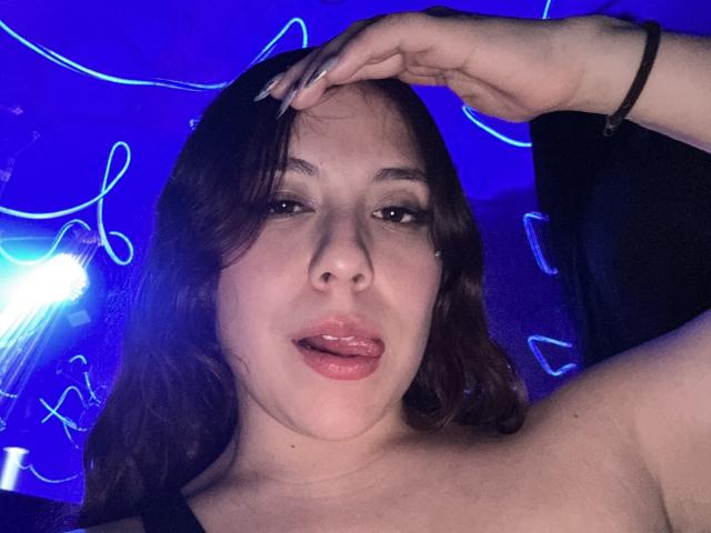 AmberlStha has dark brown hair, slightly parted lips with gloss, and a small nose piercing. She's sticking her tongue out, with a hand in her hair, wearing a black top.