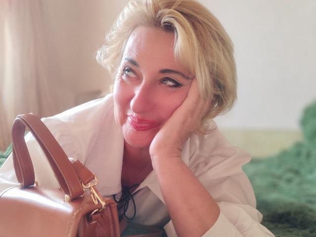 MilanaAlvis with blonde hair, red lipstick, and white shirt, resting chin on hand, smiling at brown leather handbag. Her tits are partially visible under the shirt.