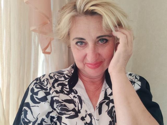 Middle-aged woman with blonde hair, blue eyes, and red lipstick, wearing a black and white floral shirt, touching her head with a hand, smiling softly at the camera.