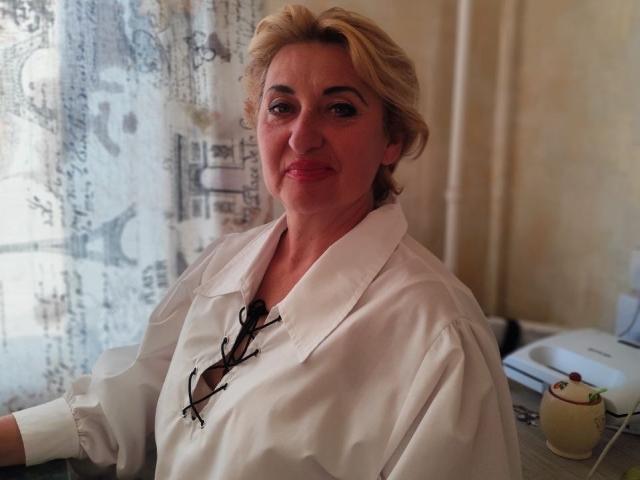 MilanaAlvis with blonde hair, wearing a white shirt, has glasses tucked in the collar, smiling, and has a cute face with pink lipstick, in a softly lit room.