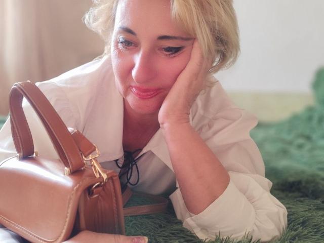 MilanaAlvis with blonde hair, white shirt, and pink lipstick, resting her face on her hand, looking at a brown leather handbag with a gold buckle.