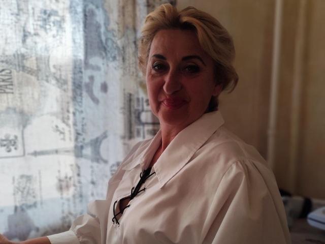 MilanaAlvis with blonde hair, wearing a white shirt, smiling softly, showing a little cleavage, and standing near a curtain with abstract patterns, looking relaxed.