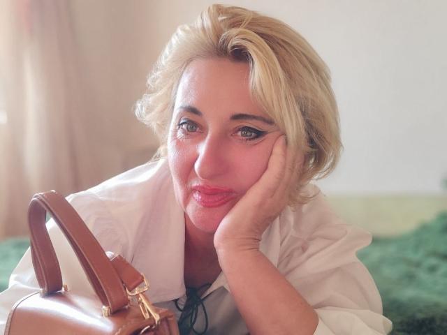 MilanaAlvis with blonde hair, pouty red lips, and green eyes, resting her face on her hand, wearing a white shirt, and holding a brown leather handbag.