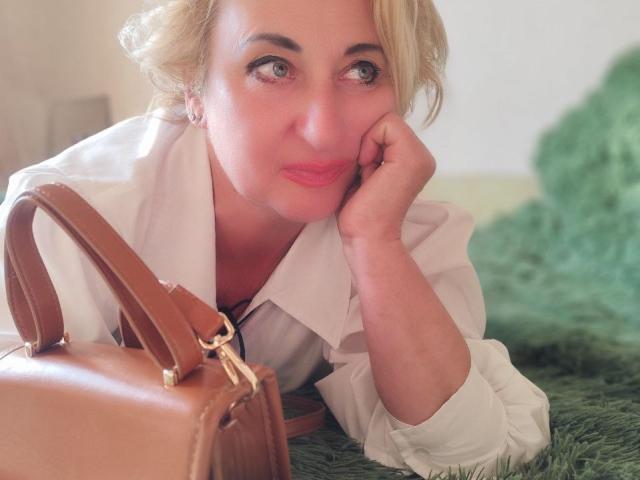 MilanaAlvis with blonde hair, lying on green carpet, wearing white shirt, brown leather purse in front, resting chin on hand, pink lipstick, looking up with soft expression.