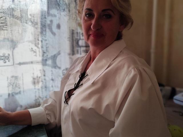 MilanaAlvis with blonde hair, wearing a white button-up shirt, black lace necklace, and a slight smile, standing by a window with lace curtains. Boobs visible under shirt.