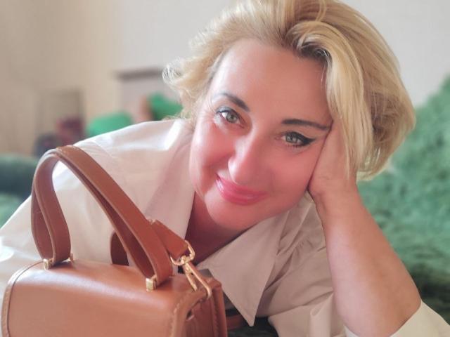 MilanaAlvis has blonde hair, green eyes, and pink lipstick, leaning on a green pillow with a brown leather handbag in front of her. She's smiling softly.