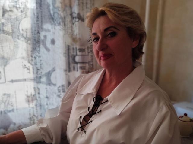 MilanaAlvis with blonde hair, wearing a white shirt with black lace-up, sitting, looking at camera, subtle smile, eyeglasses in shirt, light-skinned, medium tits, soft light, indoor setting.