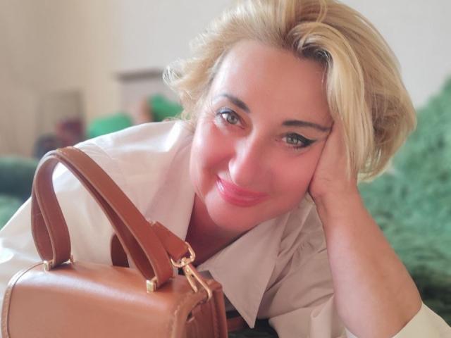 MilanaAlvis, blonde, cute, smiling, resting head on hand, wearing white shirt, brown leather purse with gold zippers, green fuzzy background, soft light, relaxed, pretty, sexy.