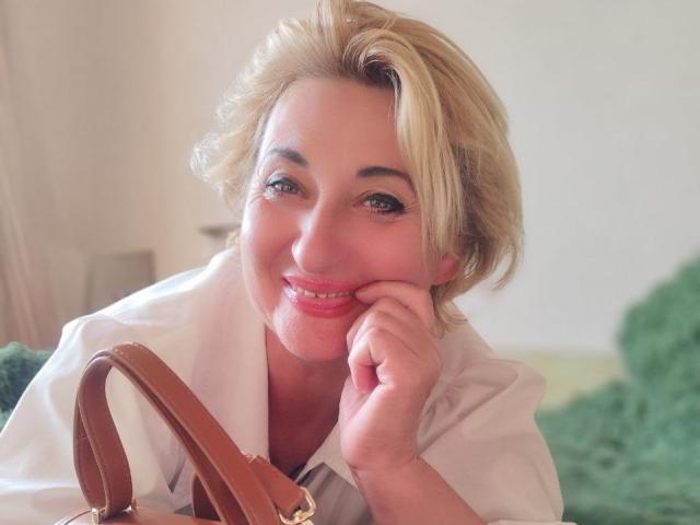 MilanaAlvis with blonde hair, smiling, wearing a white shirt, resting her chin on her hand, brown leather purse in front, green fuzzy background, soft light, pink lips.