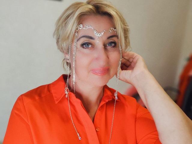 MilanaAlvis with short blonde hair, wearing an orange shirt, has a delicate silver headpiece with chains, and is touching her right ear, looking slightly smiling.