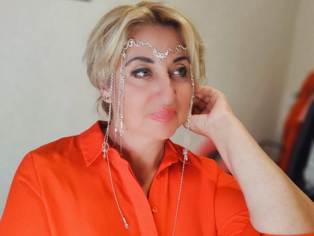 MilanaAlvis with short blonde hair, wearing an orange shirt, has a silver headpiece with chains dangling over her eyes and nose, resting her hand on her cheek.