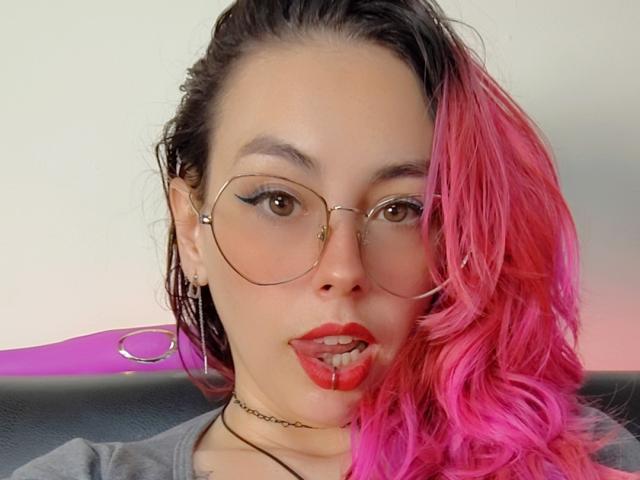 A camisa cinza de NatashaLau, com recortes, revela seus seios, seu cabelo rosa e um vibrador rosa perto de sua vulva peluda. Suas pernas estão abertas, mostrando sua vulva depilada..