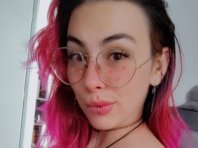 NatashaLau with pink-dyed hair, glasses, shiny lips, black choker, tattoo on shoulder, necklace, winking, shiny skin, earrings, and a cute smile, looking sexy.