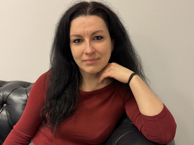 AudreyLove with wavy black hair, wearing a red long-sleeve shirt, sitting on a black leather couch, resting her chin on her hand, looking slightly annoyed.