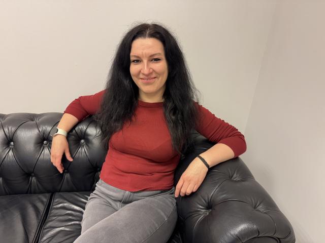 AudreyLove with long black hair, red long-sleeve top, and gray pants, sitting on a black leather couch, smiling, wearing a black wristband, and a black watch.