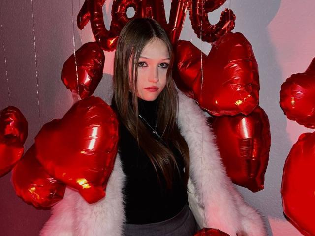 Bubochka, avec ses longs cheveux bruns, portant un pull noir à col roulé et une veste en fourrure blanche, entourée de ballons en forme de cœur rouges brillants, regarde droit devant la caméra avec une expression sérieuse..