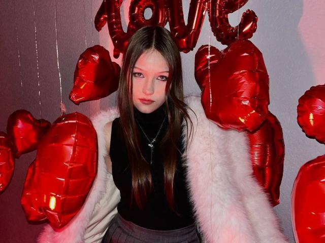 Bubochka, con cabello largo y castaño, vestida con un cuello alto negro, un abrigo de piel blanca y una falda gris, rodeada de globos rojos con forma de corazón, tiene una expresión seria y lleva un collar con una cruz..