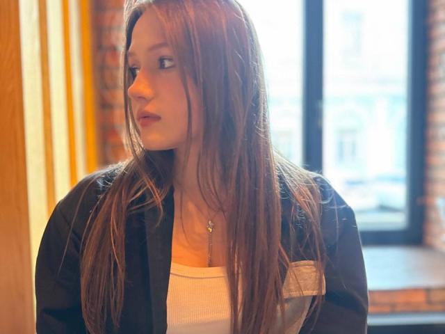 Bubochka with long brown hair, looking to the side, wearing a black jacket over a white top, small necklace, soft light on her face, window in the background.