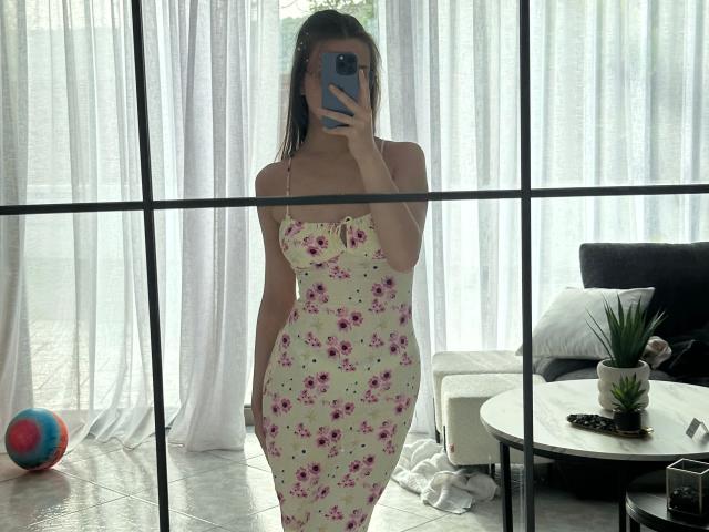 LunaLou-hot in a strapless floral dress, standing, holding a phone, covering her tits, white curtains, black-framed mirror, gray sofa, potted plant, and colorful ball in the background.