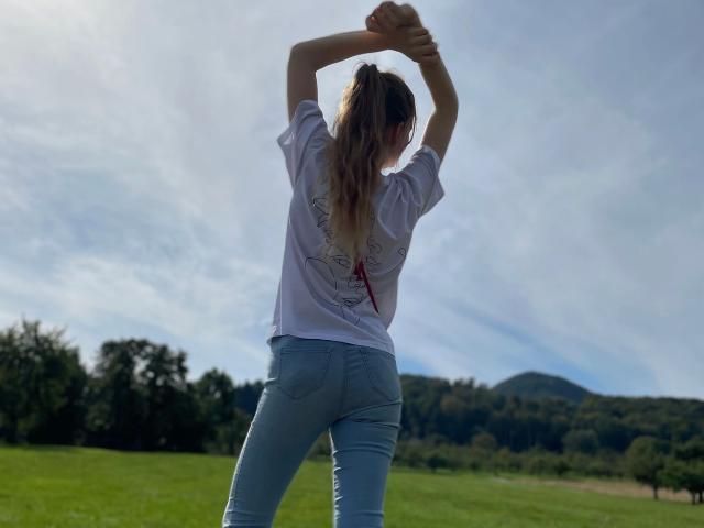 GeileNachbarin-hot with arms raised, wearing a loose white shirt and tight blue jeans, standing in a grassy field, showing her ass, under a cloudy sky.