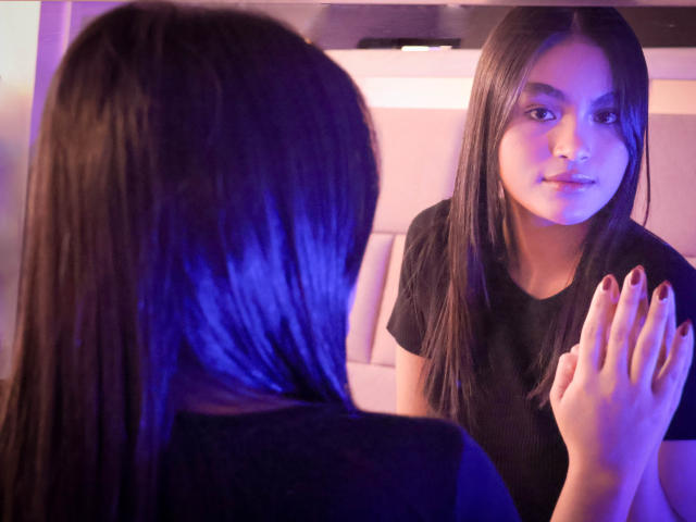 Miia Palmer, con cabello negro largo y vestida con una blusa negra, se mira en un espejo, con su mano derecha, que tiene esmalte de uñas rojo, levantada. En el reflejo, se la ve sonriendo..
