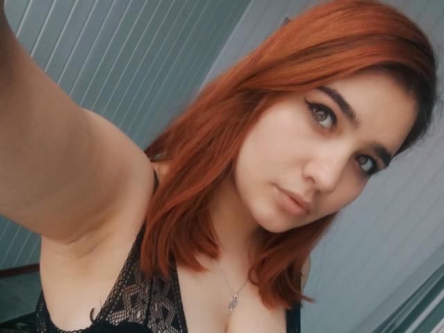 BomboMichaela has red hair, black lace top, showing her tits, and a neutral expression. She's wearing a necklace with a small pendant. Her arm is raised for the selfie.