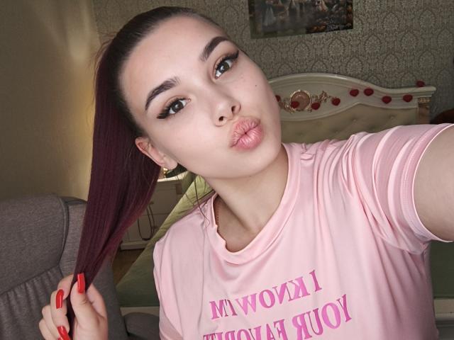 Niferttiti with puffy lips, holding her red-manicured hand in her maroon hair, wearing a pink shirt, has fair skin, green eyes, and a high-ponytail.