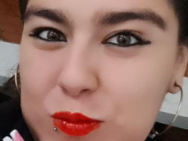 Il viso di TaylorNova, con rossetto rosso, eyeliner nero e un piercing al labbro, ha un'espressione seria e sopracciglia scure, e indossa grandi orecchini a cerchio. I suoi occhi sono leggermente rivolti di lato..