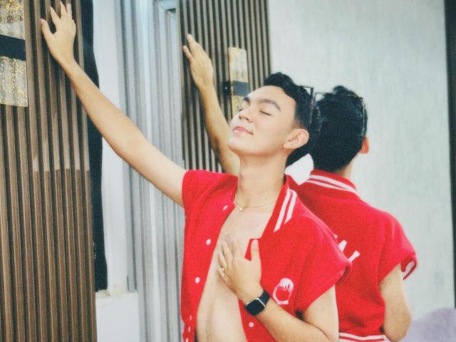 DeusBeauty with short black hair, eyes closed, smiling, wearing an open red shirt, exposing a flat chest, hand on chest, other hand on wall, reflected in mirror.