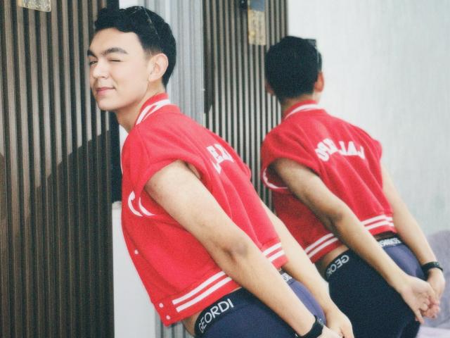 DeusBeauty, Asian guy with short black hair, red sports jacket, and blue underwear, standing with hands behind back, smiling slightly, reflection in window, daytime, casual, cute.