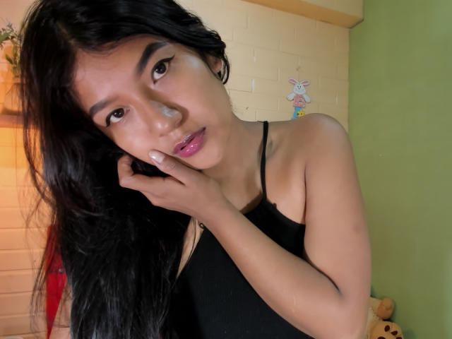 ScarletKry with long black hair, wearing a black tank top, touching her cheek, pink lips, and dark eyebrows, in a green and white room.