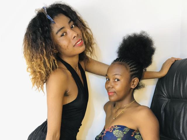 Silviesirene with curly brown hair and black halter dress, standing, arm around smaller silviesirene with afro and floral dress, black leather chair, smiling, red lipstick, gold necklace.