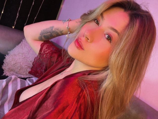 SweetAbey, con i capelli biondi, le labbra rosse e un tatuaggio sul braccio, indossa una camicetta rossa in pizzo, con il seno leggermente visibile, e posa con una mano dietro la testa..