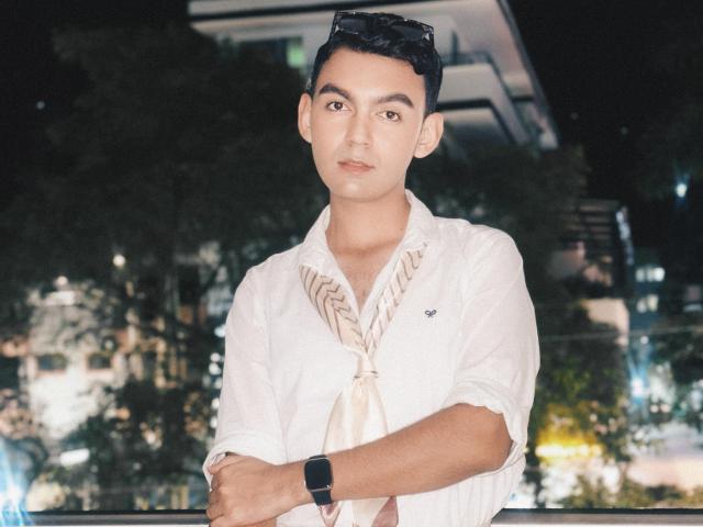 DeusBeauty, a cute Asian dude with short black hair, wearing a white shirt and beige scarf, looks serious with arms crossed, black watch on, sunglasses on head.