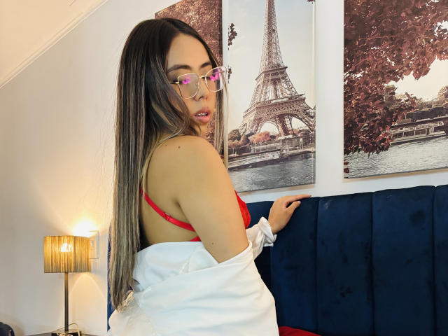 LannaKane, com cabelos castanhos longos, sutiã vermelho, camisa branca sem mangas, óculos, olhando para trás, sentada em um sofá de veludo azul, com uma obra de arte de Paris na parede, uma luminária à esquerda, luz quente..