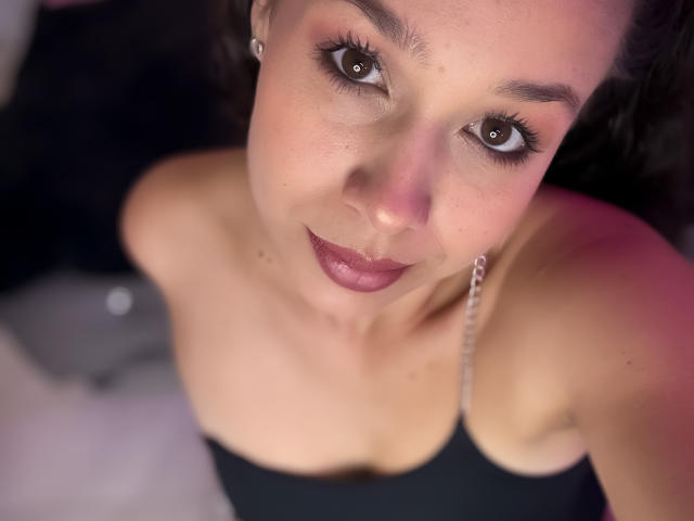 EvaMar met bruine ogen, donkerroze lippenstift en een zwart topje met bandjes, waarbij haar blote schouders en kleine borsten zichtbaar zijn, kijkt op naar de camera met een lichte glimlach..