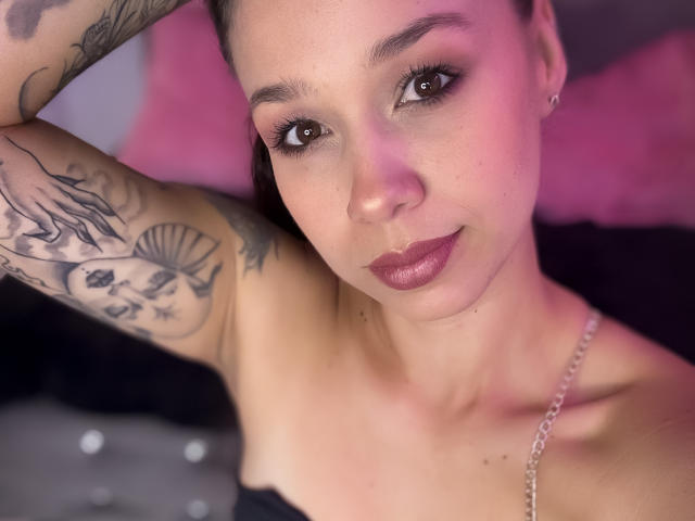 EvaMar, con ojos marrones, lápiz labial oscuro y tatuajes en el brazo, acostada, mirando a la cámara, vestida con una correa negra y una cadena de plata, con iluminación rosa..