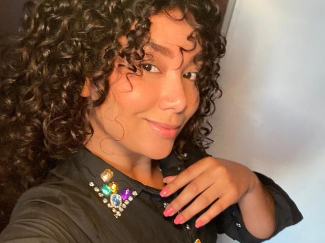Ashlley69, com cabelo preto encaracolado, pele morena e esmalte rosa, vestindo uma camisa preta com botões coloridos e brilhantes, sorrindo com um piercing no nariz e tocando o queixo..
