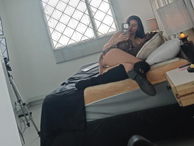 EymyFox, con i capelli scuri, una maglia di pizzo e stivali neri, sdraiata su un letto, che mostra il sedere, le gambe e il telefono, in una camera da letto con specchi, con un'attrezzatura per la videocamera..